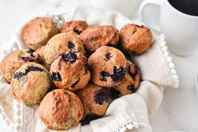 Blueberry mini muffins in a towel-lined basket next to a mug of coffee.
