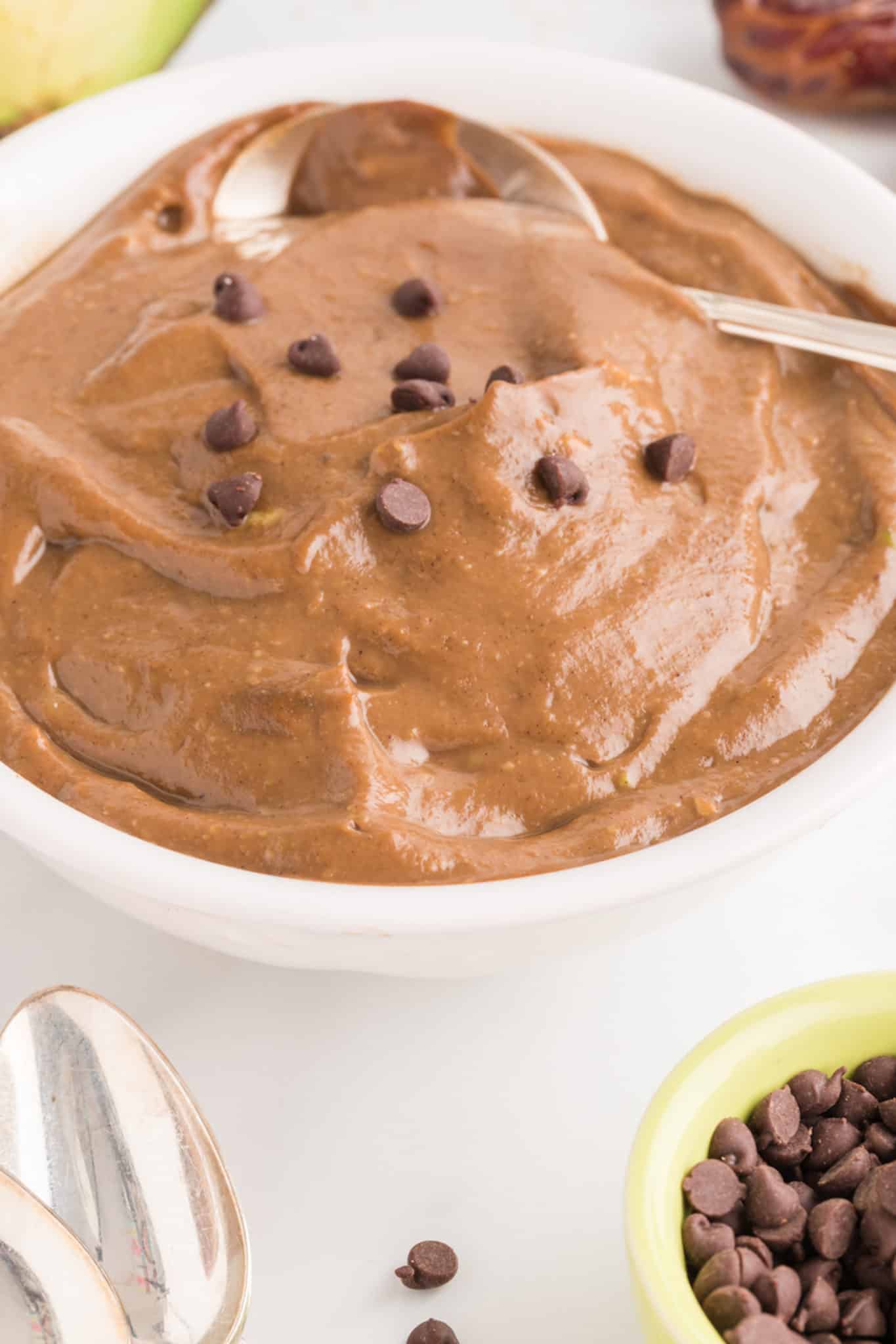 A white bowl with a spoon is filled with chocolate avocado pudding topped with chocolate chips.