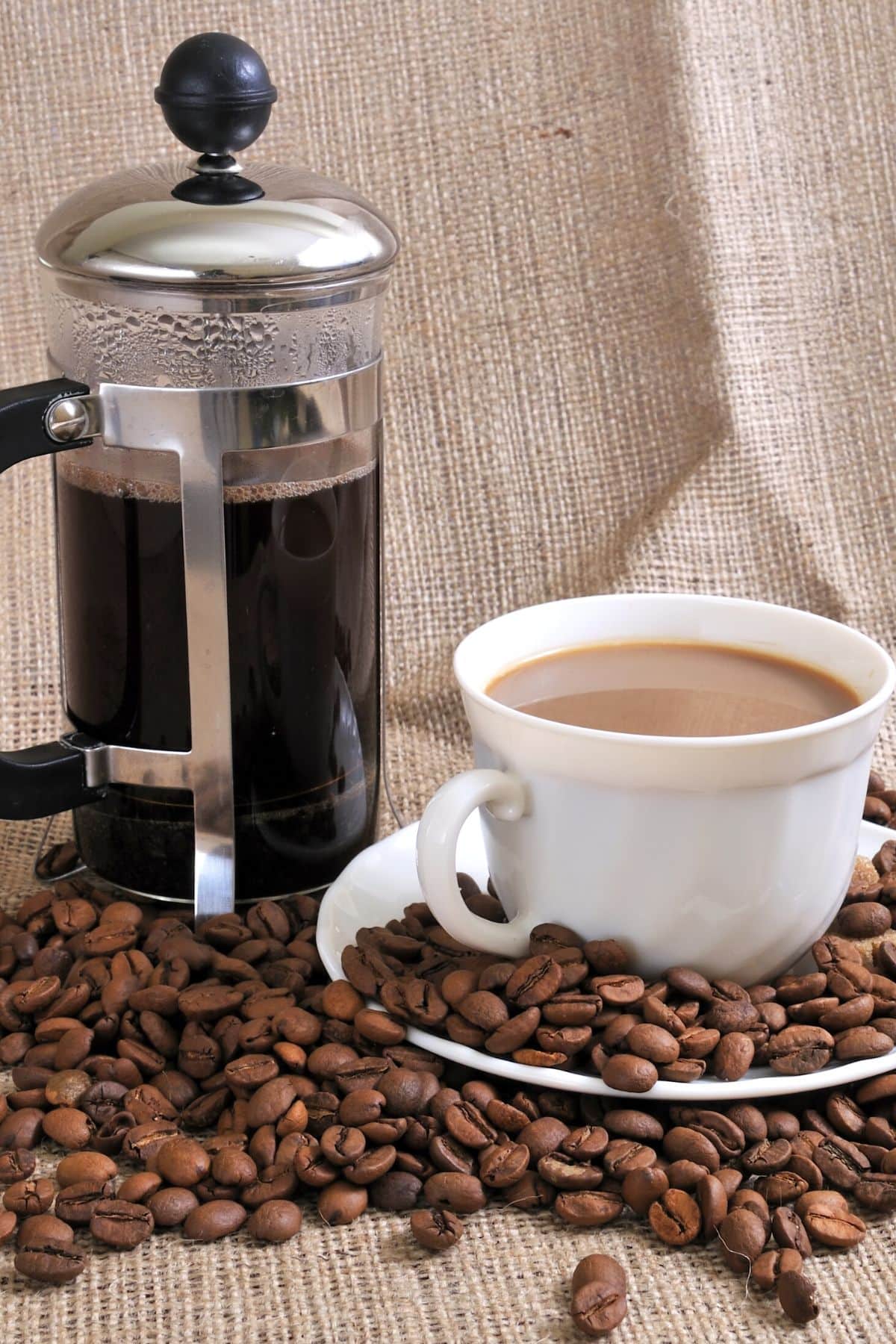 a French press and cup of coffee surrounded by coffee beans.